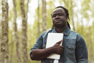 African man holding book or notebook in the park. Student studying in the nature clipart