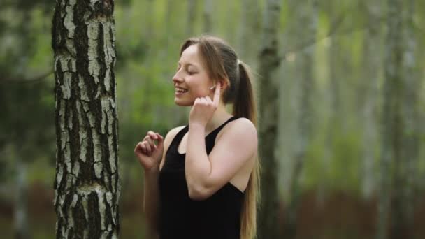 En ung kaukasisk kvinna i naturen. Lyssnar på musiken på de trådlösa hörlurarna och vidrör trädbarken. Långsamma rörelser — Stockvideo