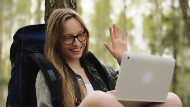 Νεαρή λευκή κοπέλα με γυαλιά και σακίδιο που αναπαύεται στη φύση και έχει βιντεοκλήση σε φορητό υπολογιστή με οικογένεια και φίλους — Αρχείο Βίντεο