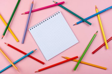 Pembe arka planda bir defter. Renkli kalemler etrafa saçılmış..