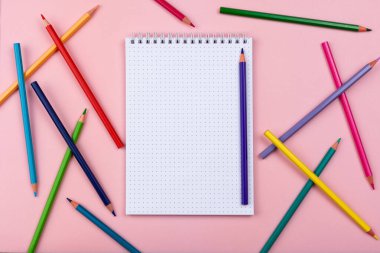 Pembe arka planda bir defter. Renkli kalemler etrafa saçılmış..
