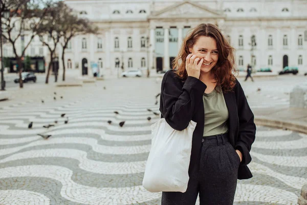 Millennial woman with canvas bag laughing at camera. Rossio Square with famous Portuguese pavement in Lisbon, Portugal