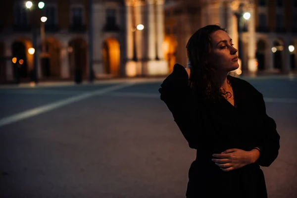 Half length portrait of young woman wearing black, standing alone in the street. Night time, street light, contemplative