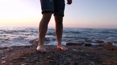 Bacağında küçük dövmesi olan genç bir çocuk gün batımında kayalarda yürüyor.