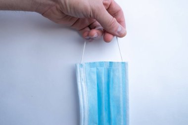 Bir adamın eli beyaz arka planda mavi, tek kullanımlık bir tıbbi maske tutuyor.