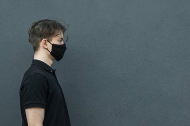 Siyah tişört ve siyah tıbbi maskeyle gezen genç adamın portresi. Virüsleri ve koronavirüs salgınını, covid-19 salgınını korumak ve durdurmak için gri arka planda. Yüzünde siyah maske olan adam. Virüs koruması. Profilde siyah tişörtlü adam var.-