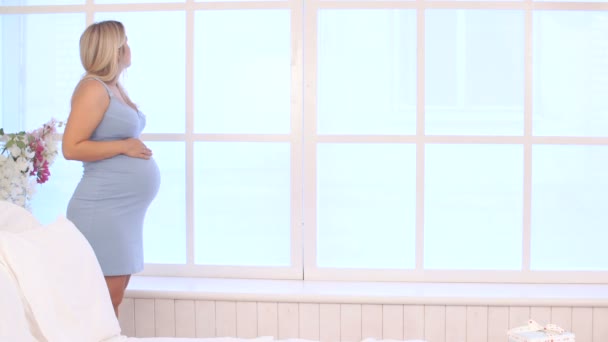 Вагітна жінка в блакитній сукні ходить уздовж вікна — стокове відео