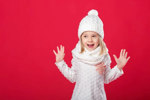 Malá usměvavá blondýnka v bílé čepici a šálu na červené pozadí — Stock fotografie