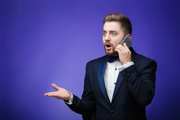 Hombre de éxito en esmoquin y pajarita hablando por teléfono. hombre de negocios sosteniendo smartphone, fondo azul —  Fotos de Stock