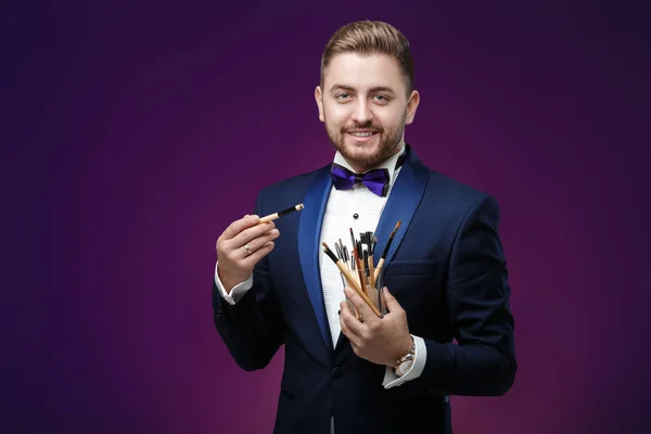 Joven con barba en esmoquin sostiene cepillo de maquillaje y sonriendo — Foto de Stock