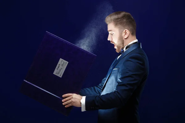 Joven con barba sosteniendo libro y luz mágica sobre fondo oscuro — Foto de Stock