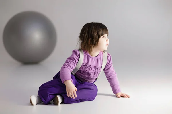 Kleines Mädchen in lila Kleidung mit großem Ball für Fitness auf grau — Stockfoto