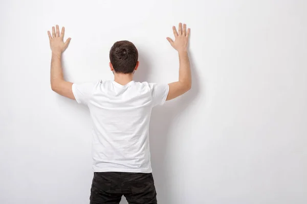 Νεαρός άνδρας στο λευκό t-shirt με τα χέρια ψηλά κοντά στον τοίχο, πίσω όψη. δ — Φωτογραφία Αρχείου