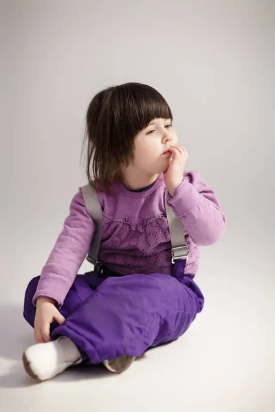 Kleines nettes Mädchen in lila Kleidung denkt auf grauem Hintergrund — Stockfoto