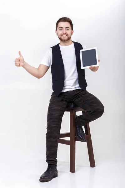 Joven hombre guapo con barba en camisa blanca y chaleco negro sosteniendo tableta y mostrando el pulgar hacia arriba signo sobre fondo gris — Foto de Stock