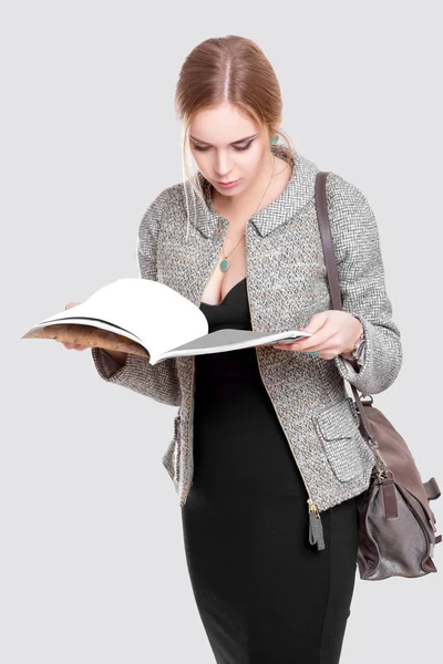 Hermosa mujer de negocios rubia en vestido negro, chaqueta leyendo una revista sobre fondo gris —  Fotos de Stock