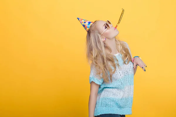Porträtt av roliga blond kvinna i födelsedag hatt och blå skjorta på gul bakgrund. Fest och party. — Stockfoto