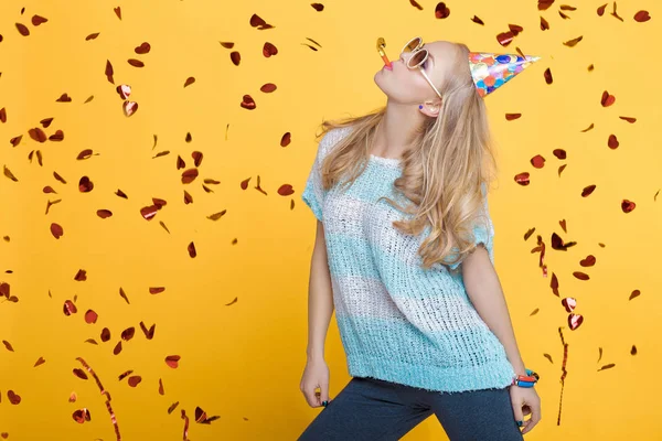 Portrait de femme blonde drôle en chapeau d'anniversaire et confettis rouges sur fond jaune. Célébration et fête . — Photo