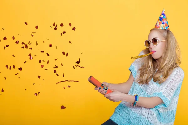 Porträtt av roliga blond kvinna i födelsedag hatt och röd konfetti på gul bakgrund. Fest och party. — Stockfoto