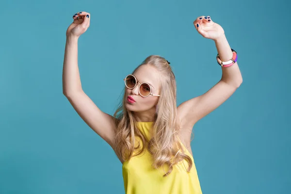 Portrett av en vakker blond kvinne i solbriller og en gul skjorte som danser på blå bakgrunn. Carefree summer . – stockfoto