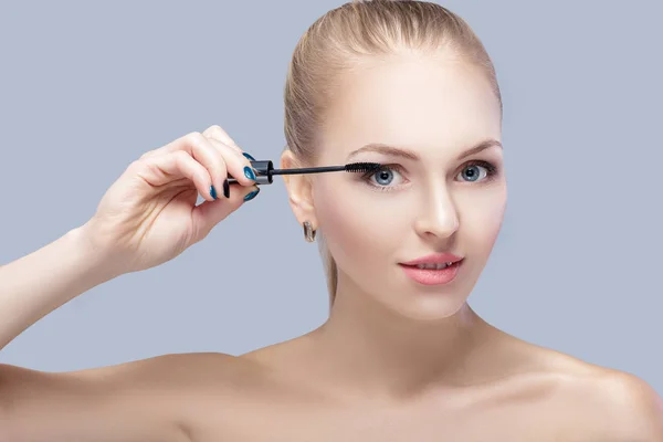 Bela mulher loira aplicando maquiagem no rosto em fundo cinza. maquiagem perfeita. escova de rímel — Fotografia de Stock