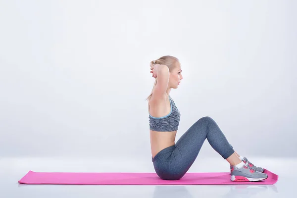 Bela menina loira no sportswear faz exercícios sobre fitness Mat no fundo cinza — Fotografia de Stock