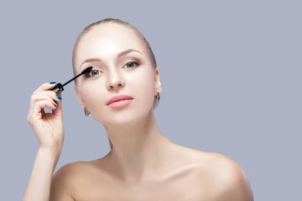 Bella donna bionda applicare il trucco sul viso su sfondo grigio. trucco perfetto. spazzola di mascara — Foto Stock