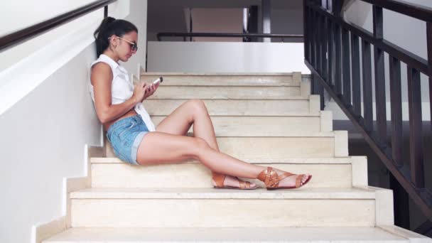 Junge brünette Frau in kurzen Hosen, Sonnenbrille auf der Treppe sitzend und im Sommer telefonierend. — Stockvideo