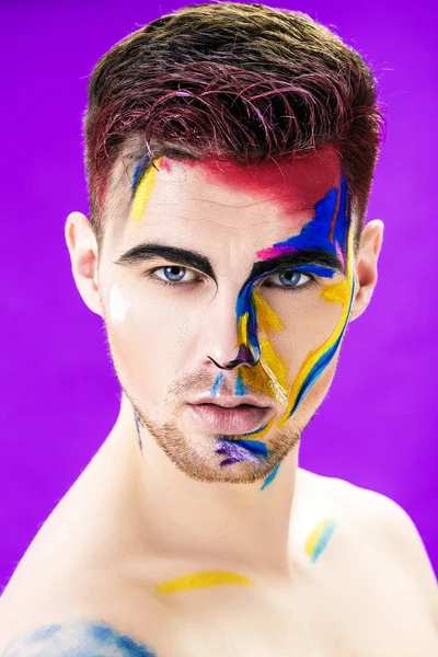 Retrato de hombre joven atractivo con pintura facial de color sobre fondo púrpura. Moda de maquillaje profesional. maquillaje de arte de fantasía — Foto de Stock