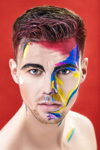 Retrato de jovem atraente homem com pintura facial colorida sobre fundo vermelho. Moda de Maquiagem Profissional. maquiagem arte extravagante — Fotografia de Stock