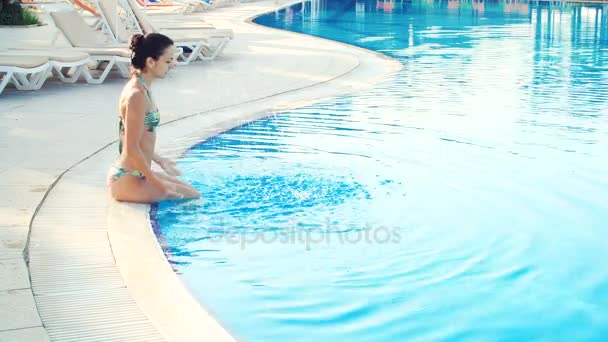 Joven morena mujer en bikini y gafas de sol sentado en la orilla de la piscina y hace chapoteo en el día de verano soleado — Vídeo de stock