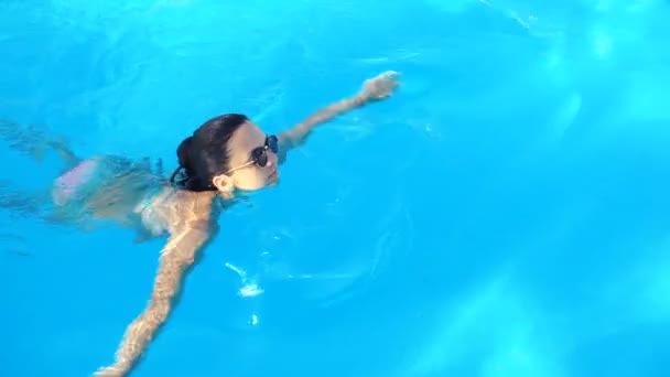 Joven morena en bikini y gafas de sol nada en la piscina el soleado día de verano — Vídeos de Stock