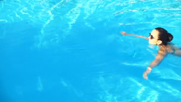 Joven morena en bikini y gafas de sol nada en la piscina el soleado día de verano — Vídeos de Stock