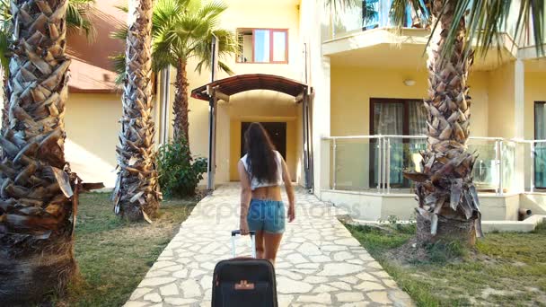 Молодая брюнетка в шортах и каблуках поставляется с чемоданом между пальмами в отеле в летнее время. отдых и отдых — стоковое видео