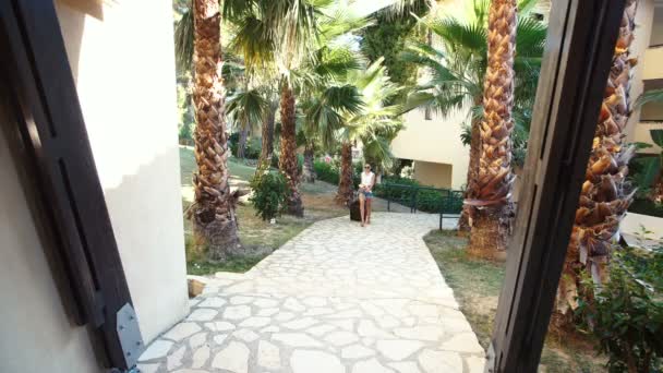 Genç esmer kadın şort, güneş gözlüğü ve topuklu bavulla otel palmiye ağaçları arasında geliyor. tatil — Stok video