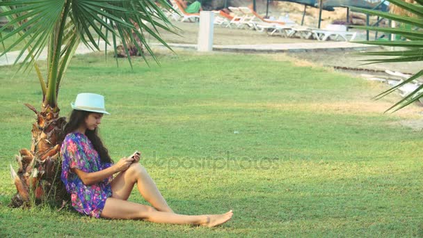 Молода засмагла жінка з довгим волоссям, що сидить під тропічними пальмами влітку. вона використовує смартфон — стокове відео