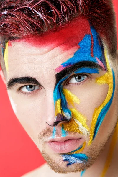 Retrato de hombre joven con pintura facial de color sobre fondo rojo. Moda de maquillaje profesional. fantasía arte maquillaje — Foto de Stock