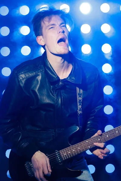 Jovem músico de rock tocando guitarra elétrica e cantando. Estrela de rock no fundo dos holofotes — Fotografia de Stock