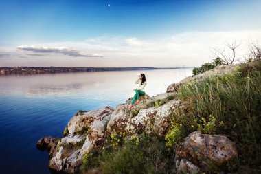 Genç kadın rock üzerinde oturur ve gün batımında nehir manzarasına sahiptir