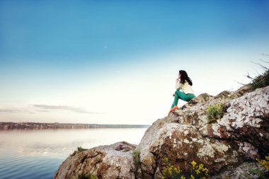 Genç kadın rock üzerinde oturur ve gün batımında nehir manzarasına sahiptir