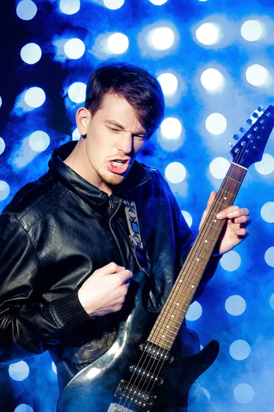 Jovem músico de rock atraente tocando guitarra elétrica e cantando. Estrela de rock no fundo dos holofotes — Fotografia de Stock