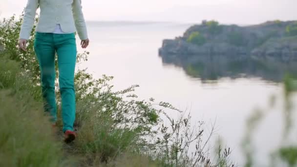 Chica en pantalones verdes camina por el camino a lo largo del río a las flores — Vídeo de stock