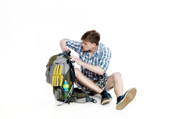Young tourist is sitting and looking in backpack something on white background. traveler is preparing for hike — Stock Photo, Image