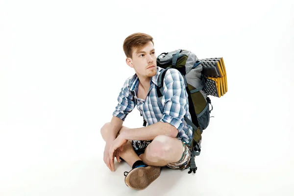 Joven turista está sentado con la mochila sobre fondo blanco. viajero se está preparando para la caminata — Foto de Stock