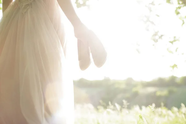 Novia en vestido de novia sostiene los zapatos contra el sol. fotografía de arte — Foto de Stock