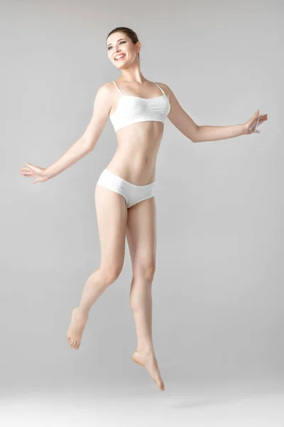 Тонка красива жінка з ідеальним тілом у білій білизні стрибає на сірому фоні — стокове фото