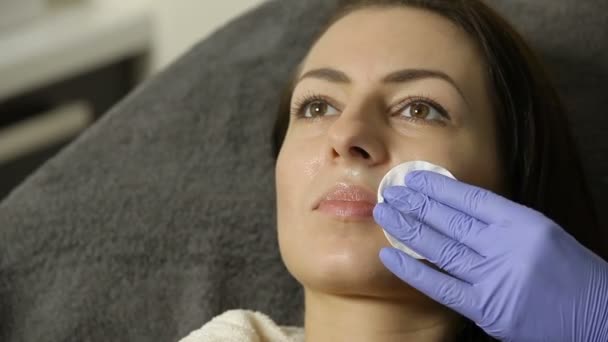 Αισθητικός σκουπίζει το πρόσωπό της γυναίκας ασθενούς με βαμβάκι σφουγγάρι πριν από τη διαδικασία. απολύμανση — Αρχείο Βίντεο