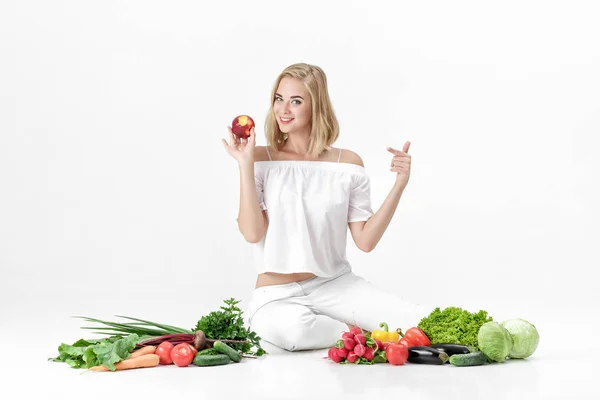 Hermosa mujer rubia en ropa blanca y un montón de verduras frescas sobre fondo blanco. chica está comiendo nectarina — Foto de Stock