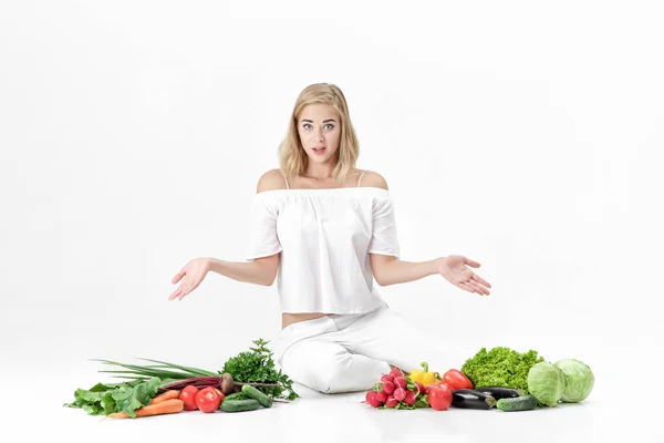 Femme blonde frustrée écarte les mains dans les deux sens et beaucoup de légumes frais sur fond blanc — Photo