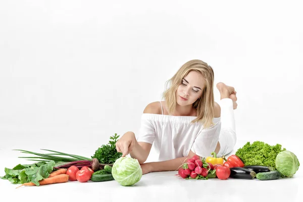 Belle femme blonde en vêtements blancs et beaucoup de légumes frais sur fond blanc — Photo